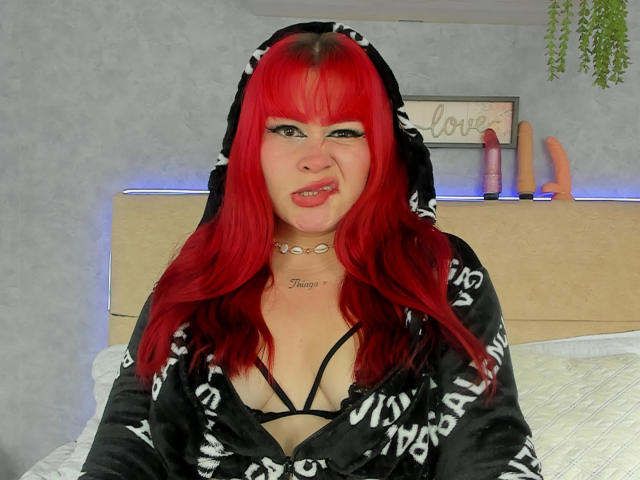 KendraVelvet with bright red hair, black and white hooded robe, black strappy bra, tongue out, choker, tattoos, bed, and blue-lit sex toys in the background.
