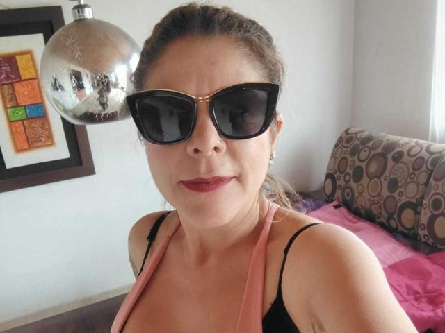 MartinaFiore mit großen schwarzen Sonnenbrillen, einem pinken Halstopp und braunen Haaren im Pferdeschwanz, die ihre Brüste zur Schau stellt und roten Lippenstift trägt, steht in der Nähe einer silbernen Discokugel..