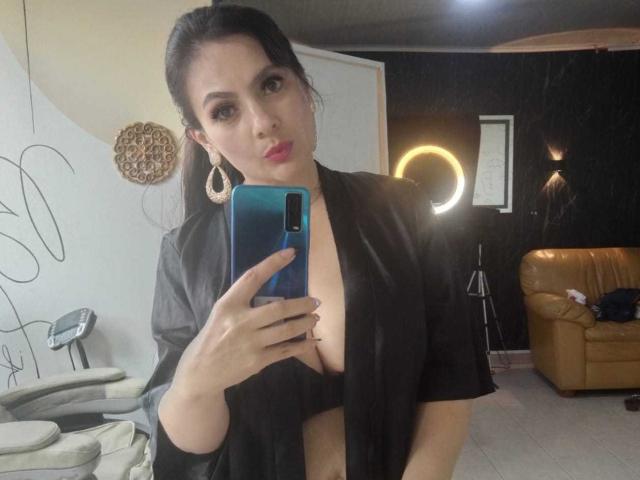 JakyKidman with dark hair, red lipstick, and hoop earrings, taking a mirror selfie in a black robe, exposing her tits, holding a blue phone, in a modern room.