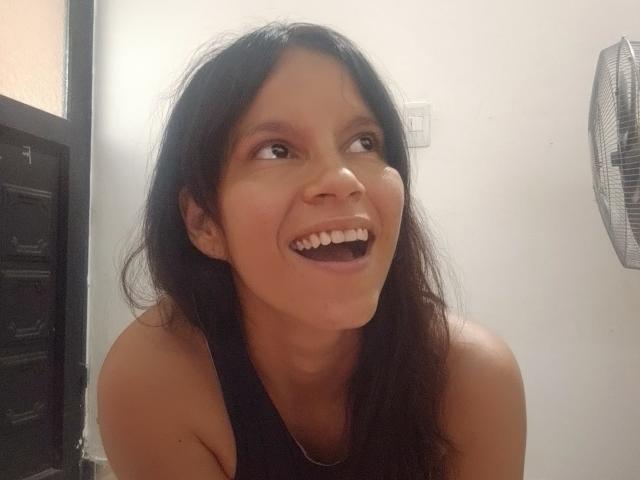 EstrellaFandy with black hair, smiling widely, looking up with her mouth open, showing her teeth. She's wearing a black tank top, and a fan is on the right.