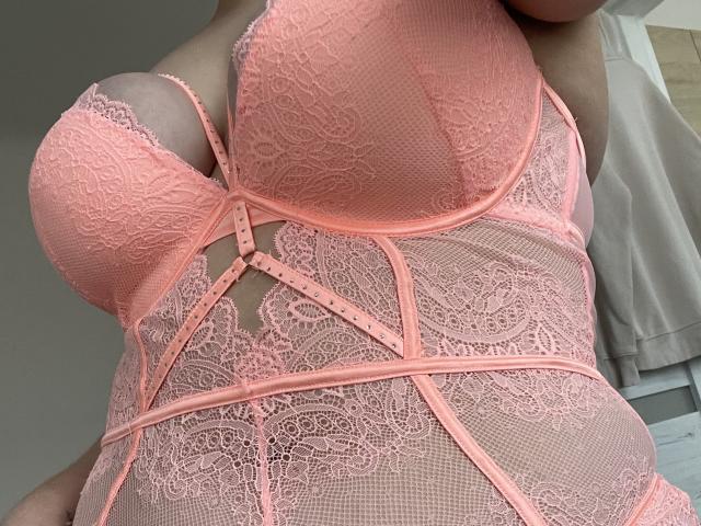LilyyMoon wearing a pink lace lingerie set with her big boobs prominently displayed, lace patterns covering her belly and chest, straps and buttons adding to the design.
