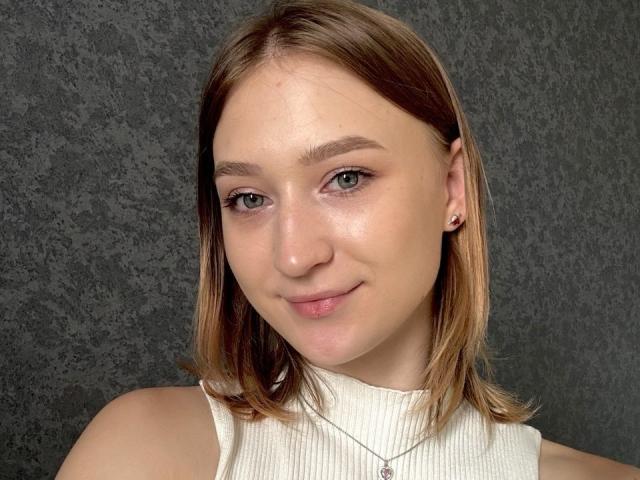 LilySweetBabe has light green eyes, a small nose, and a subtle smile. She's wearing a white sleeveless top, with shoulder-length brown hair and a silver necklace.