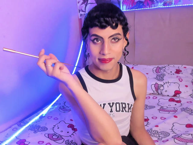 EimyFlorian with dark curly hair, red lipstick, and white sleeveless shirt holding a makeup brush, sitting on a Hello Kitty bed, in a purple-lit room.