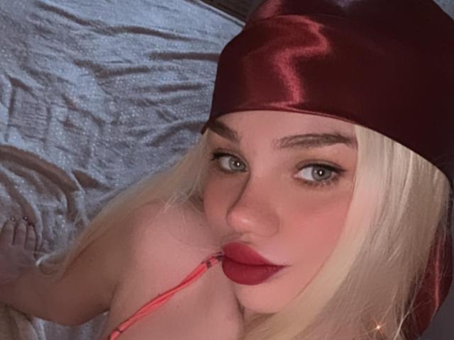 DianaRhonda with long blonde hair, red satin hat, and red lipstick, biting a pink strap, has blue eyes, and is looking up at the camera, lying on a bed.