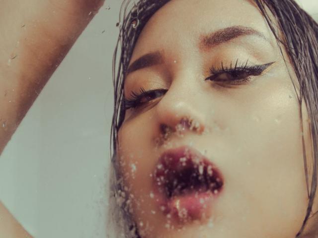 LiannaHalls' face with water droplets, eyes half-open, long black eyelashes, slightly parted pink lips, wet brown hair, and a relaxed expression. Her nose is centered in the image.