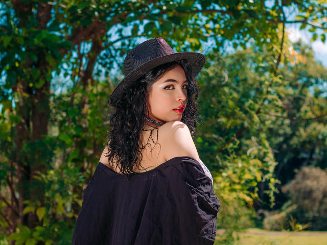 MeghanRider with curly black hair, wearing a black hat and off-shoulder black top, looking over her shoulder, red lipstick, green leafy background, sunny day, showing her bare shoulders.