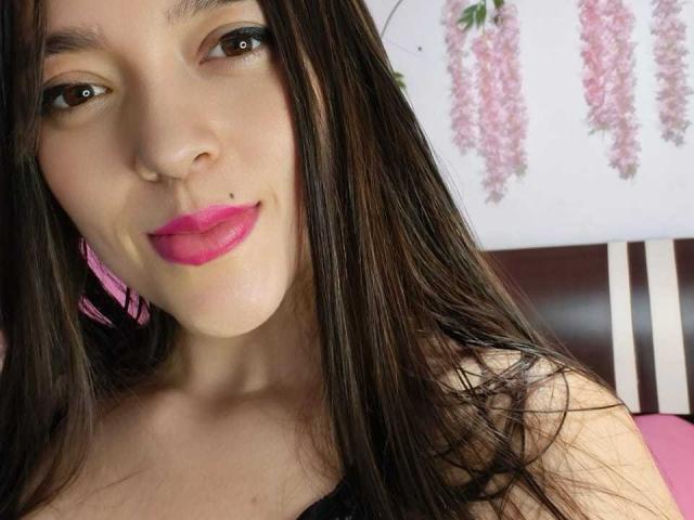 LucyMaurer has long brown hair, pink lipstick, and a small black beauty mark on her upper lip. She's smiling with pink earrings visible. Her tits are not shown.