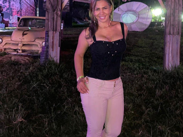 BrigiteWalsh with tan skin, black strapless top showing her tits, white pants, smiling, long brown hair, holding umbrella, standing in front of vintage car, grassy background.