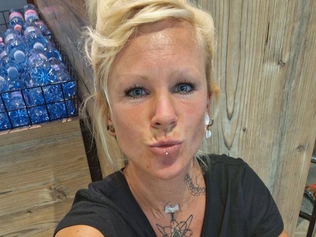 Rojia-hot has blonde hair, blue eyes, black shirt, pierced nose, and pouty lips making a kissy face. Visible tattoos on neck and chest. Water bottles in background.