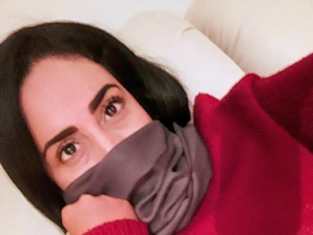 MaturesSugar69 with dark hair, red sweater, and gray mask covering lower face, looking up with brown eyes, resting hand on chin. Soft, warm lighting.