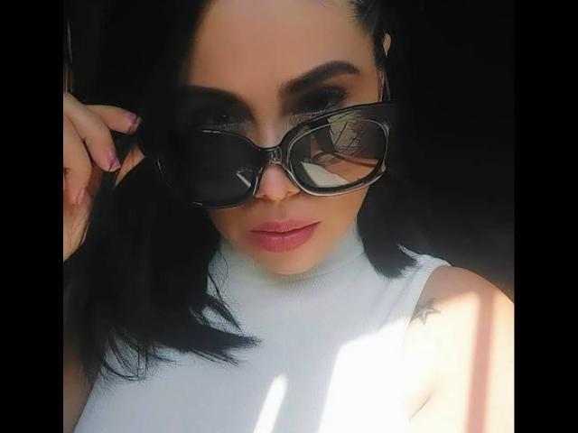 MaturesSugar69 with black hair, wearing oversized black sunglasses, white turtleneck, pink lipstick, and a tattoo on the left shoulder, holding her hair. Boobs slightly visible under turtleneck.