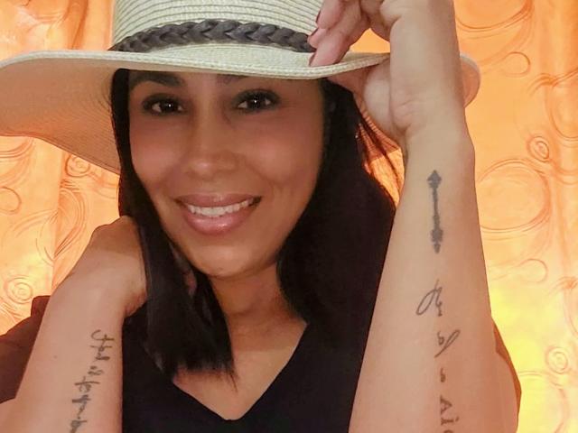 MaturesSugar69 with tan skin, dark hair, smiling, wearing a straw hat, black top, holding hat, showing tattoos on right forearm (arrow) and left forearm (cursive letters).