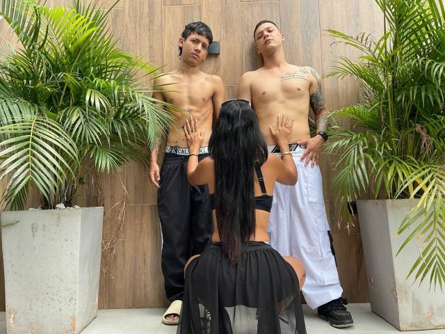 Dos hombres sin camisa de pie, una mujer llamada TheMansionDeep con cabello negro y piel bronceada, frente a ellos, con las manos sobre los abdominales de los hombres, vestida con lencería negra, una toalla blanca, pantalones negros, y plantas en macetas alrededor..