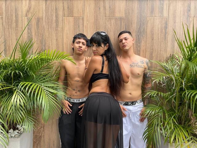 Trois personnes debout : TheMansionDeep avec de longs cheveux noirs, un soutien-gorge noir et une jupe noire, et deux hommes sans chemise en sous-vêtements Calvin Klein, entourés de plantes vertes..