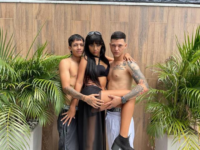 Three shirtless guys with tattoos holding TheMansionDeep up by her ass. She's wearing a black sheer dress, long black hair, and sunglasses on her head. Palm plants in foreground.