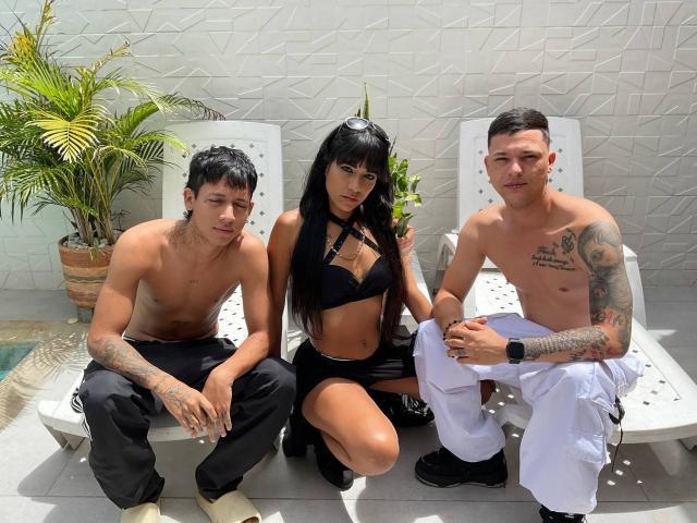 Tres personas sentadas en sillas blancas, TheMansionDeep con cabello negro largo, un pequeño sostén negro y pantalones cortos, flanqueada por dos hombres sin camisa con tatuajes, pantalones negros y pantalones blancos..