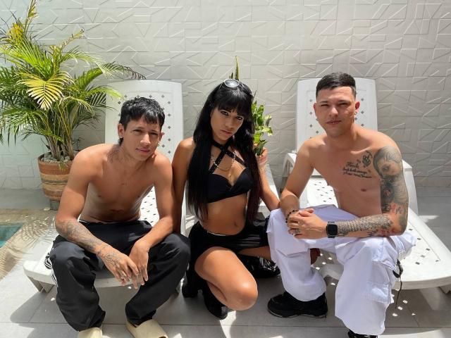 Trois personnes assises sur des chaises blanches : TheMansionDeep porte un bikini noir et a de longs cheveux noirs, deux hommes sans chemise portant des pantalons noirs et blancs, tous deux tatoués et avec des cheveux courts..