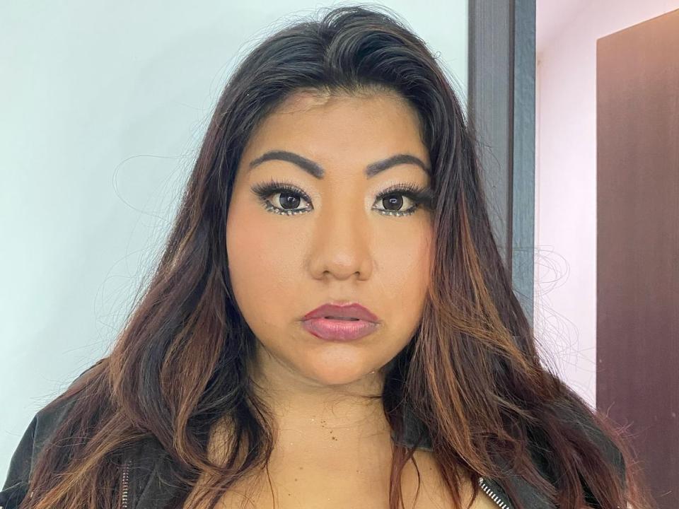 SashaPalmeraa has long, wavy brown hair, thick eyebrows, and full pink lips with subtle makeup. She's wearing a black top, looking directly at the camera with a neutral expression.