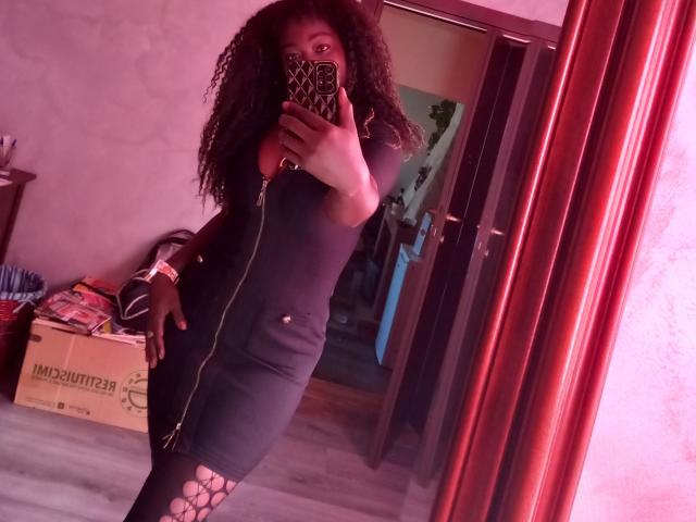 Ajova mit lockigem Haar, einem schwarzen, hochgeschlossenen Kleid und Leggings mit Ausschnitten, macht ein Selfie vor einem Spiegel und zeigt dabei ihre dunkelbraune Haut und ihre mittelgroße Brust. Sie hält ein Handy mit einer Handyhülle mit Leopardenmuster..