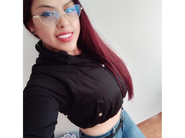ElaiaaBakerr with red hair, wearing glasses, black crop top, showing her small tits, and blue jeans, smiling, standing in a room with wooden floors.