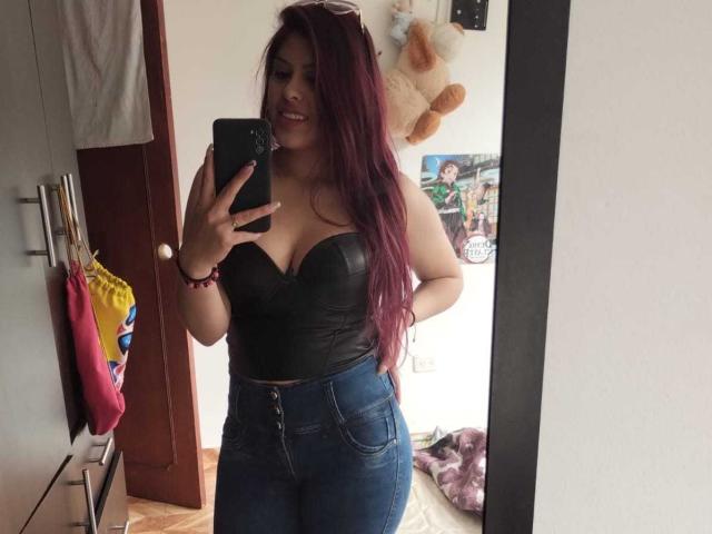 ElaiaaBakerr with red hair, black strapless leather top, showing big tits, and tight blue jeans, taking a mirror selfie, smiling, holding a phone, in a bedroom.
