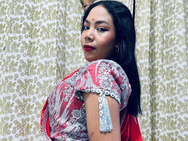 KalindaSajanii with dark hair, red lipstick, and a red and silver patterned outfit, showing her ass with a tassel, looking over her shoulder, smiling.