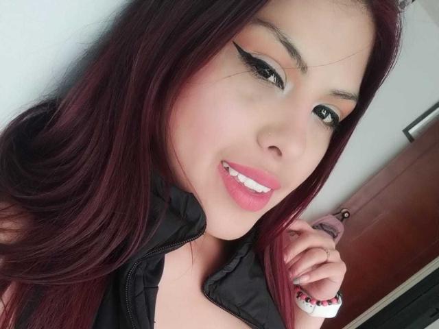 ElaiaaBakerr tem cabelo ruivo, delineador preto, batom rosa e está sorrindo, mostrando dentes brancos. Ela está vestindo uma jaqueta preta, e sua mão tem contas vermelhas e brancas..
