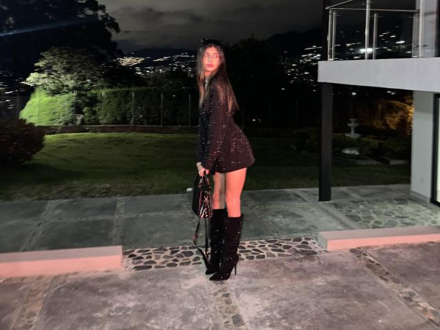 CiaSweet, avec de longs cheveux bruns, une robe noire pailletée, des bottes hautes noires, tenant un sac en cuir noir, debout sur une terrasse en pierre la nuit, avec des yeux brillants, sensuelle..