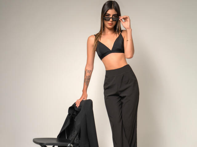 CiaSweet, avec de longs cheveux bruns, portant un soutien-gorge noir et un pantalon taille haute, tenant une veste noire, avec des tatouages sur le bras droit, des lunettes noires, debout, la main près des lunettes, souriante..