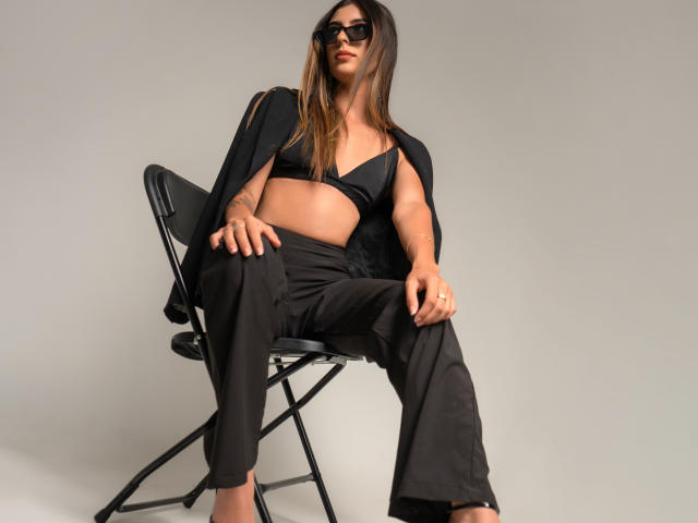 CiaSweet, con cabello largo y castaño, gafas de sol negras, un sostén negro, pantalones negros y una chaqueta negra, sentada en una silla negra, mostrando un tatuaje en su mano derecha..