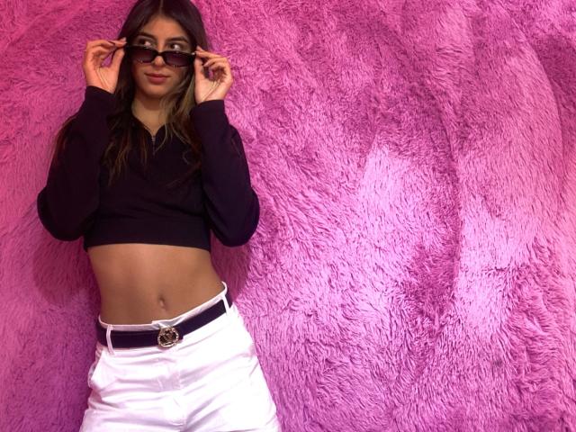 CiaSweet mit langen, braunen Haaren, die ein schwarzes Crop-Top und weiße Hosen mit einem lila Gürtel trägt, hält eine schwarze Sonnenbrille vor ihr Gesicht und zeigt so ihr flaches, trainiertes Gesäß..