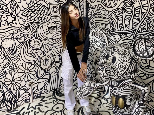 CiaSweet, avec de longs cheveux bruns, un crop top noir, un pantalon blanc et des baskets blanches, se tient près d'un mur à rayures zébrées, souriante, les mains sur les genoux. Ses seins sont petits..