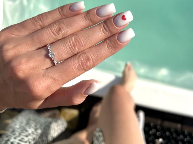 Karish's hand with white, almond-shaped nails, small red heart on index nail, silver ring, and sunlit skin, resting near her bare feet.