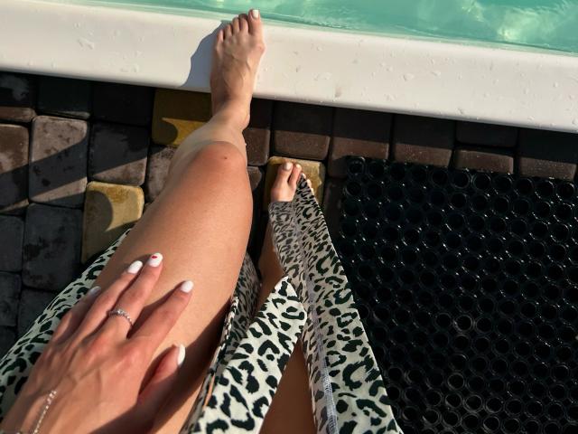 Karish's legs with a leopard print dress, one foot in a black hole-patterned mat, the other on a white pool edge. Her hand with white nails and a ring.