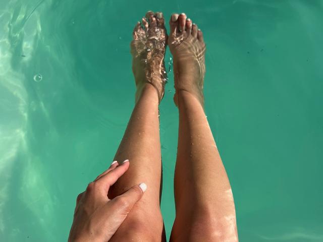 Karish's tanned legs with water droplets on her feet, her hand gripping her right thigh, toes slightly curled, standing in a clear blue-green pool.