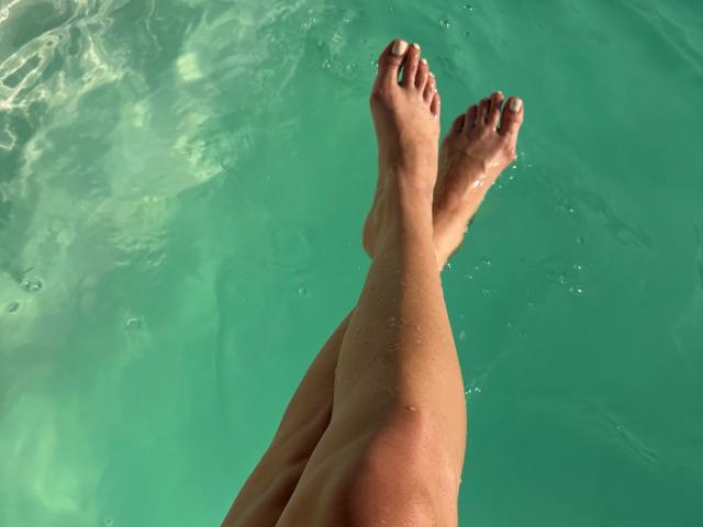 Karish's wet, tan, crossed legs with toes pointed upwards in clear green water, showing droplets on skin. Karish's feet are the focus.