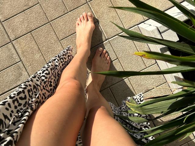 Karish's bare feet with painted toenails are crossed on a leopard-print cloth, sunlight highlighting her pale skin. Green plant leaves partially frame the bottom right.