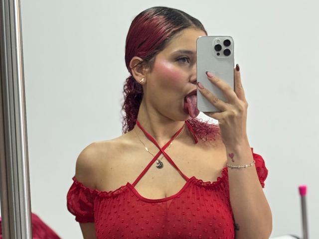 ValeryScot with red hair, tongue out, wearing a red off-shoulder top, holding a phone, taking a mirror selfie, showing her tits and red criss-cross necklace.