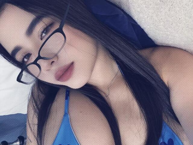 LianaBernocchi with long black hair, wearing glasses and a blue bra, lying down, showing her boobs, looking at the camera with a slight pout.