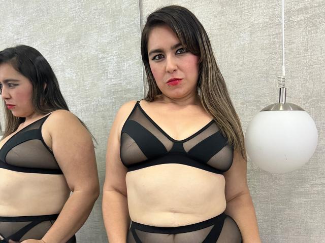TaylorNova with medium brown skin, long brown hair, red lipstick, wearing black sheer bra and panty set, showing her tits and ass, standing in front of mirror, lamp right.