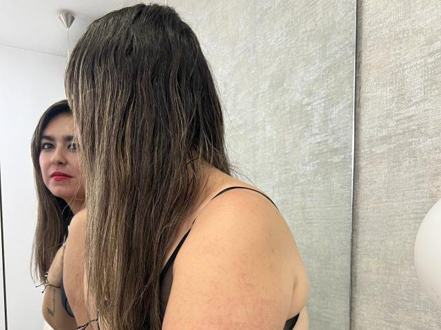 TaylorNova with long brown hair, black strap, looking at mirror, showing her boobs and a tattoo on her arm, in a bright room with white and gray walls.