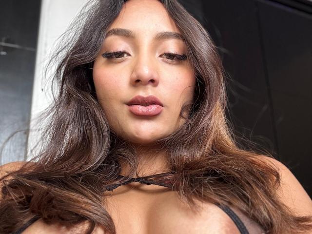 DianaFernandezz has long, wavy brown hair, full pink lips, and is wearing a black strap top, showing her tits. Her eyes are half-open, and she looks cute.