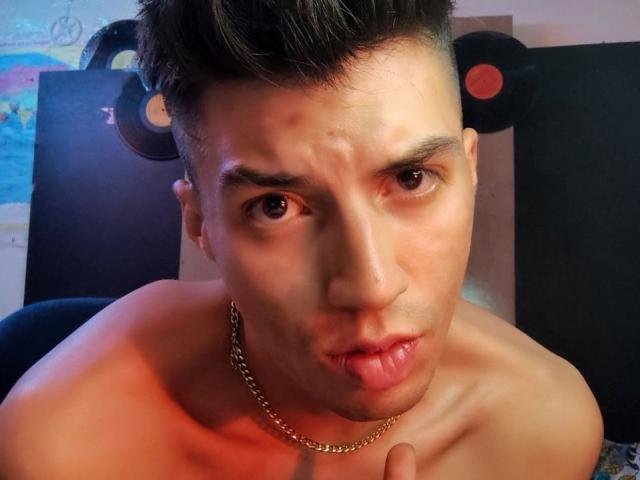 ApoloCarter, shirtless, with short dark hair, pouty lips, chain necklace, and a slight frown, has a cute, slightly annoyed expression. Vinyl records are in the background.
