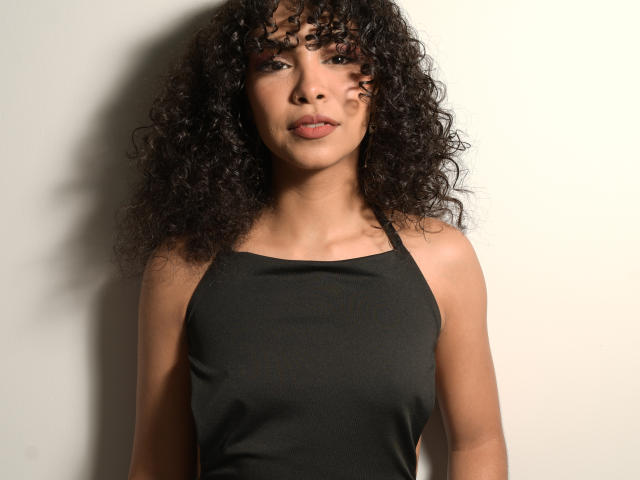 JadeMiranda with curly black hair, wearing a black halter top, has small tits and a cute face, standing against a white wall, looking slightly to the side.