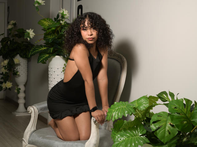 JadeMiranda, with curly black hair, kneels on a grey chair, wearing a black, backless dress, showing her small ass and slender, toned body, surrounded by green plants.