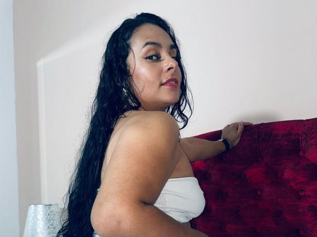 LunaSoler with long black wet hair, white tube top, red velvet couch, looking back with a slight smile, showing her tits, soft light on her tan skin.