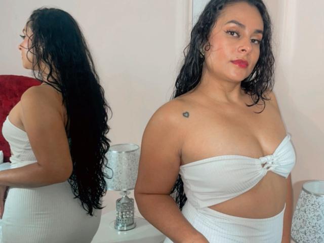 LunaSoler, con cabello negro y rizado, lleva un top blanco sin tirantes que revela pequeños senos y un corte en la cintura, está de pie junto a un espejo que refleja su trasero, con un fondo de pared blanca y una silla roja..