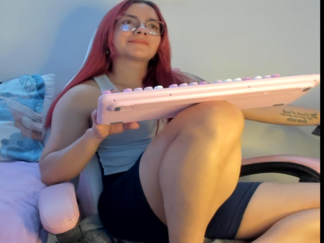 EimmyHoskX with red hair, glasses, and a white tank top, holding a pink electronic keyboard, sitting in a chair with black shorts, showing a tattoo on her arm.