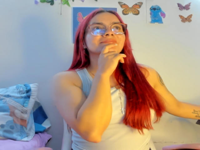EimmyHoskX with red hair, glasses, white tank top, thinking with finger on chin, light skin, small tits, tattoo on right arm, sitting on bed with blue pillow.