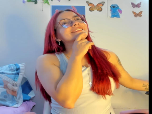 EimmyHoskX with red hair, glasses, and small tits, smiling, hand near chin, wearing a white tank top, in a brightly lit room, with butterflies on the wall.